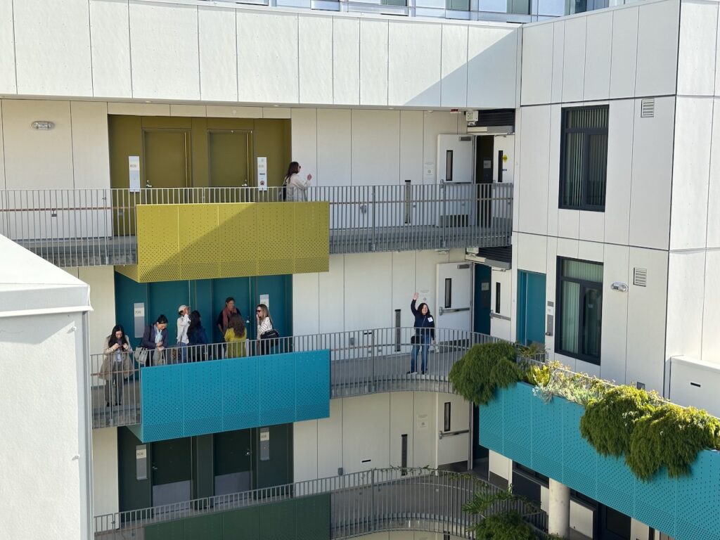 Members of the Women’s Development Collaborative standing on the balcony walkways of The Kelsey Civic Center, a modern, multi-story apartment building. On the middle floor, several members talk near a bright blue balcony while a woman further down waves at the camera. The building has bright yellow, blue, and dark green painted balconies, with green plants hanging over the balcony railings.