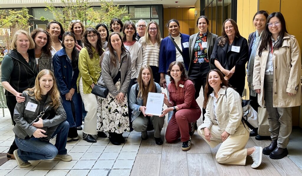 Members of the Women’s Development Collaborative, a diverse group of women in casual clothing, smile together for a photo in the courtyard at The Kelsey Civic Center, a modern apartment building. Windows, trees, and yellow walls are visible behind them. Some women stand while others kneel in front of them. Two of the women kneeling hold a small sign that reads, “Welcome!” with the Women’s Development Collaborative logo on it.