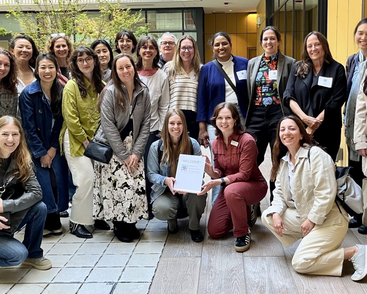 Members of the Women’s Development Collaborative, a diverse group of women in casual clothing, smile together for a photo in the courtyard at The Kelsey Civic Center, a modern apartment building. Windows, trees, and yellow walls are visible behind them. Some women stand while others kneel in front of them. Two of the women kneeling hold a small sign that reads, “Welcome!” with the Women’s Development Collaborative logo on it.