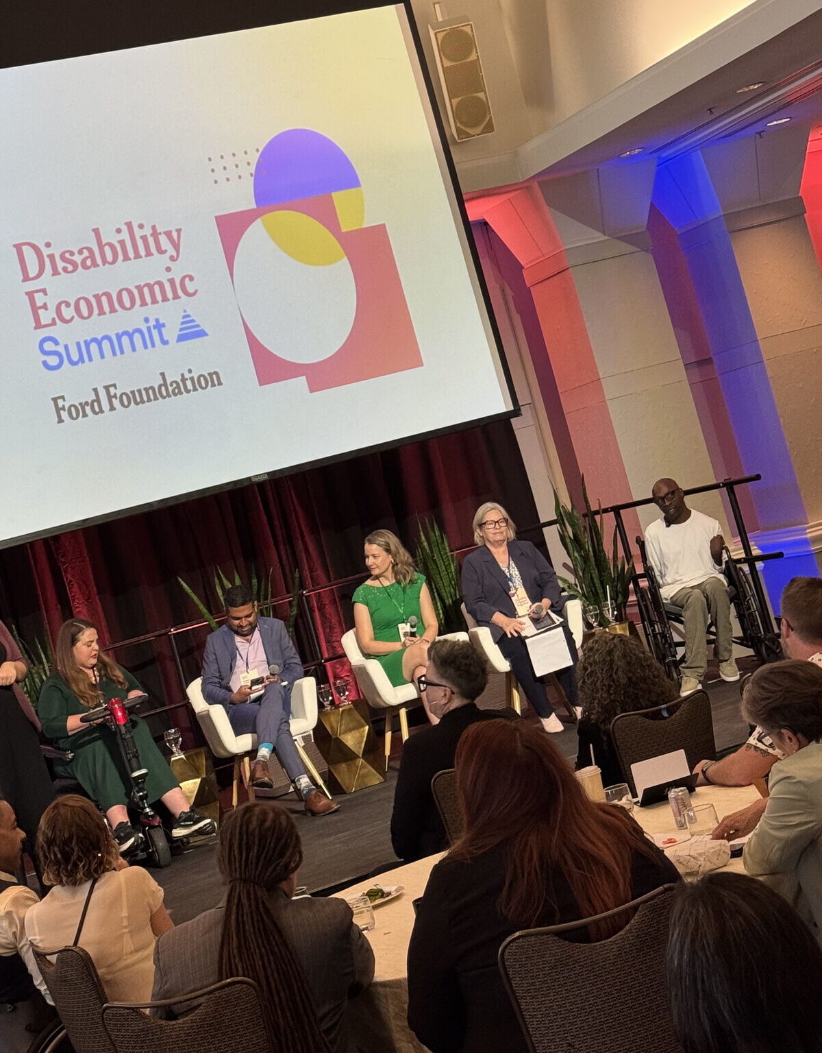 A group of panelists, with and without disabilities, of different genders and races, along with a sign language interpreter, is on a small stage. Audience members sitting around tables watch the panelists. Behind the panelists, a large projector screen displays a sign that says “Disability Economic Summit. Ford Foundation.” To the right of the text, there is a graphic design with red, purple, and yellow circles, squares, and asymmetric shapes overlapping.