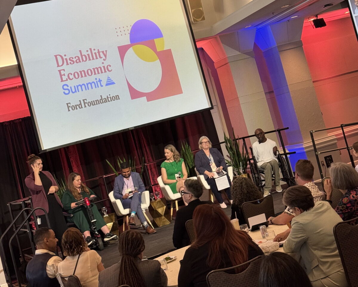 A group of panelists, with and without disabilities, of different genders and races, along with a sign language interpreter, is on a small stage. Audience members sitting around tables watch the panelists. Behind the panelists, a large projector screen displays a sign that says “Disability Economic Summit. Ford Foundation.” To the right of the text, there is a graphic design with red, purple, and yellow circles, squares, and asymmetric shapes overlapping.