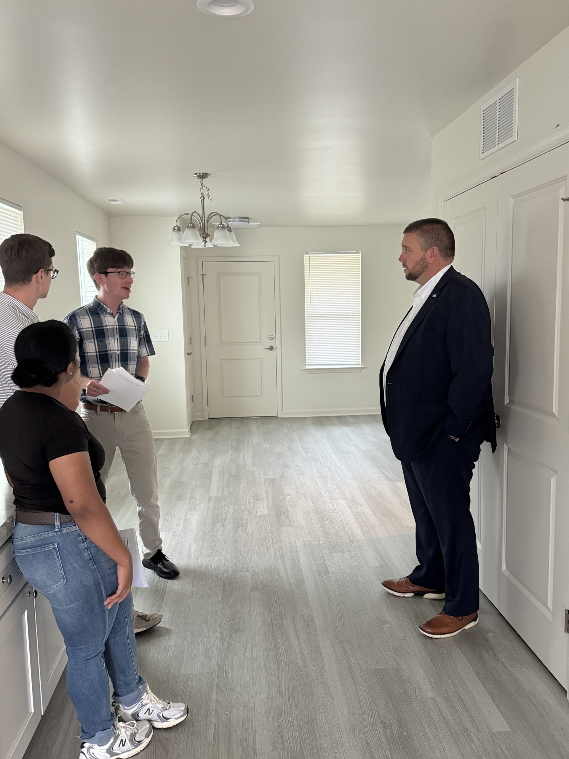 Three people give another person a tour of an empty house. One person with short brown hair, wearing a plaid shirt, a brown belt, and beige slacks, holds papers and speaks while facing the person receiving the tour. The person on the tour wears a navy suit and listens intently as the tour guide speaks.