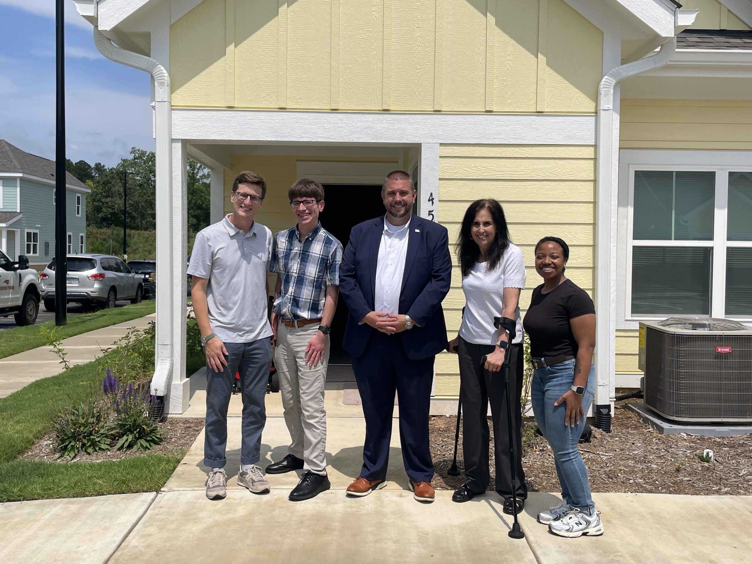 Five people stand in front of a yellow house with white trimmings. They wear a range of casual and business clothing, with some wearing sneakers, slacks, and collared shirts, and the person in the middle wearing a navy suit. The person to the right of the middle uses forearm crutches.