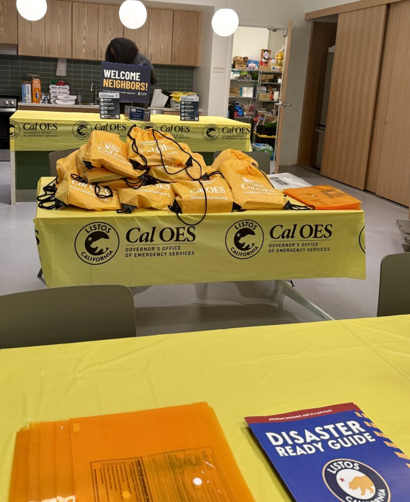 Tables covered with bright yellow tablecloths in The Kelsey Civic Center community room. The tablecloths have the California Governor’s Office of Emergency Services’ “Listos California” campaign logo on them. The logo is a black circle with a bear outline in the middle and a single star above it. The text “Listos California” is also in the circle. A disaster-ready guide pamphlet is in the foreground of the picture on a table, and yellow string backpacks are piled on another table behind it. The community room’s kitchen is visible in the background, with a person setting up in it.