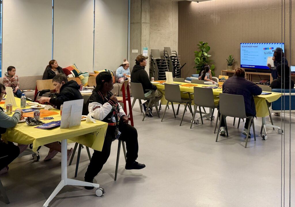 One of The Kelsey’s Inclusion Concierge stands at the front of a small community room with couches, tables, and chairs, and presents a scavenger hunt activity for the group. A slideshow is projected on a TV next to the Inclusion Concierge.