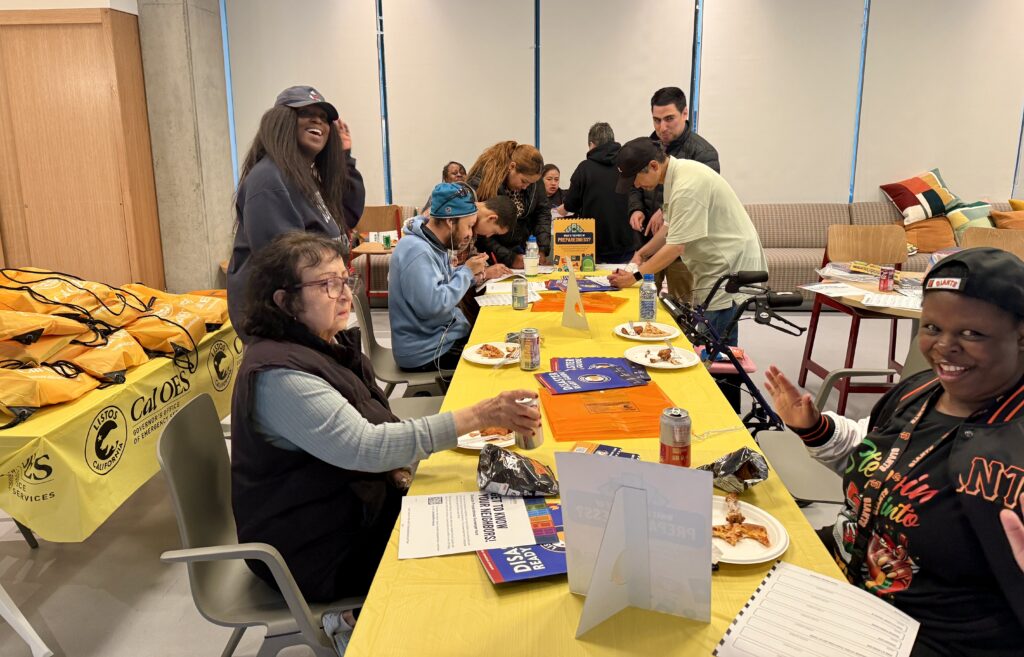 Residents of The Kelsey Civic Center sit and stand around long tables with bright yellow table cloths, pizza, drinks, and disaster preparedness handouts on them. Some are focused on writing while others smile at the camera.