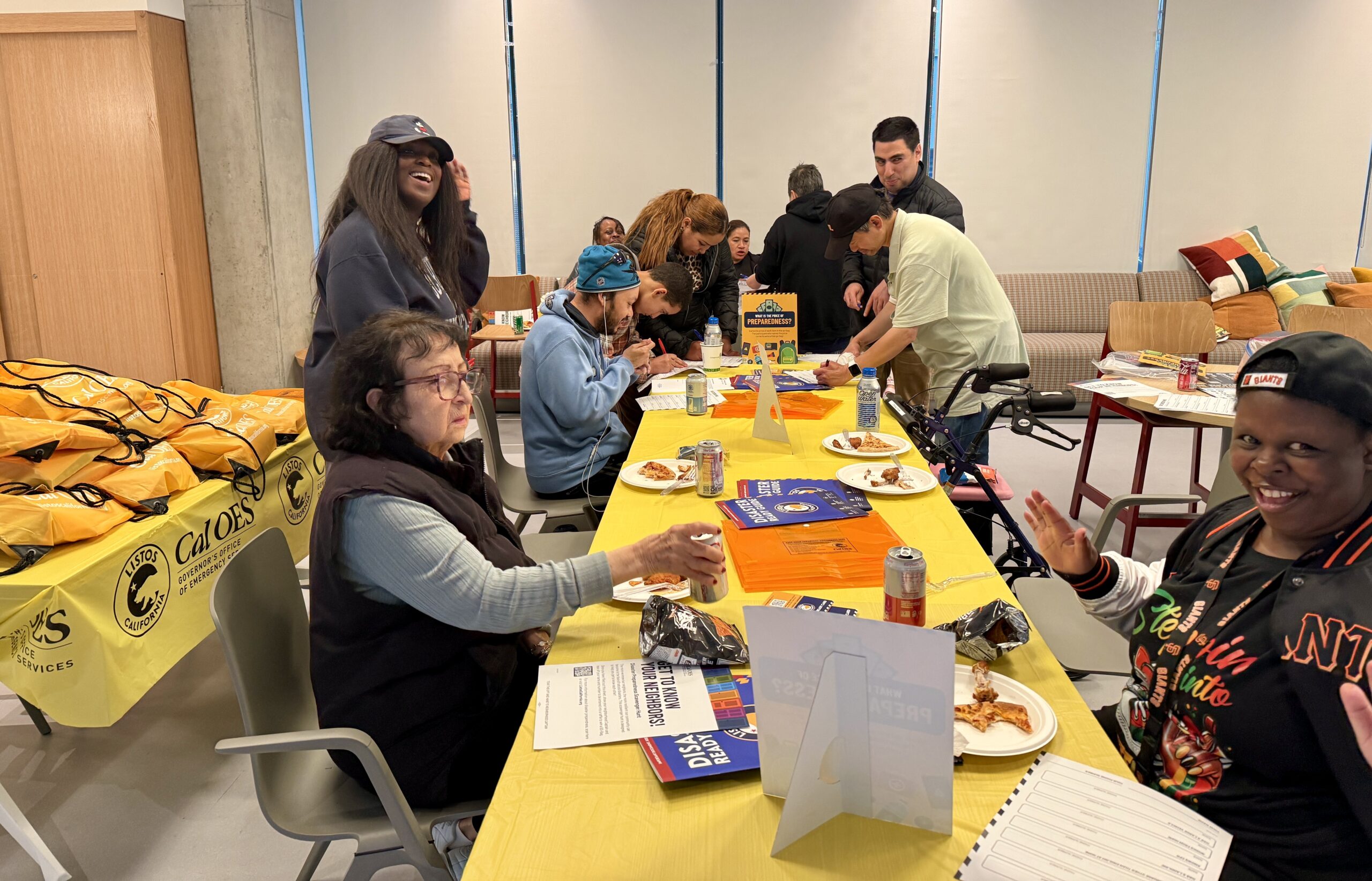 Residents of The Kelsey Civic Center sit and stand around long tables with bright yellow table cloths, pizza, drinks, and disaster preparedness handouts on them. Some are focused on writing while others smile at the camera.