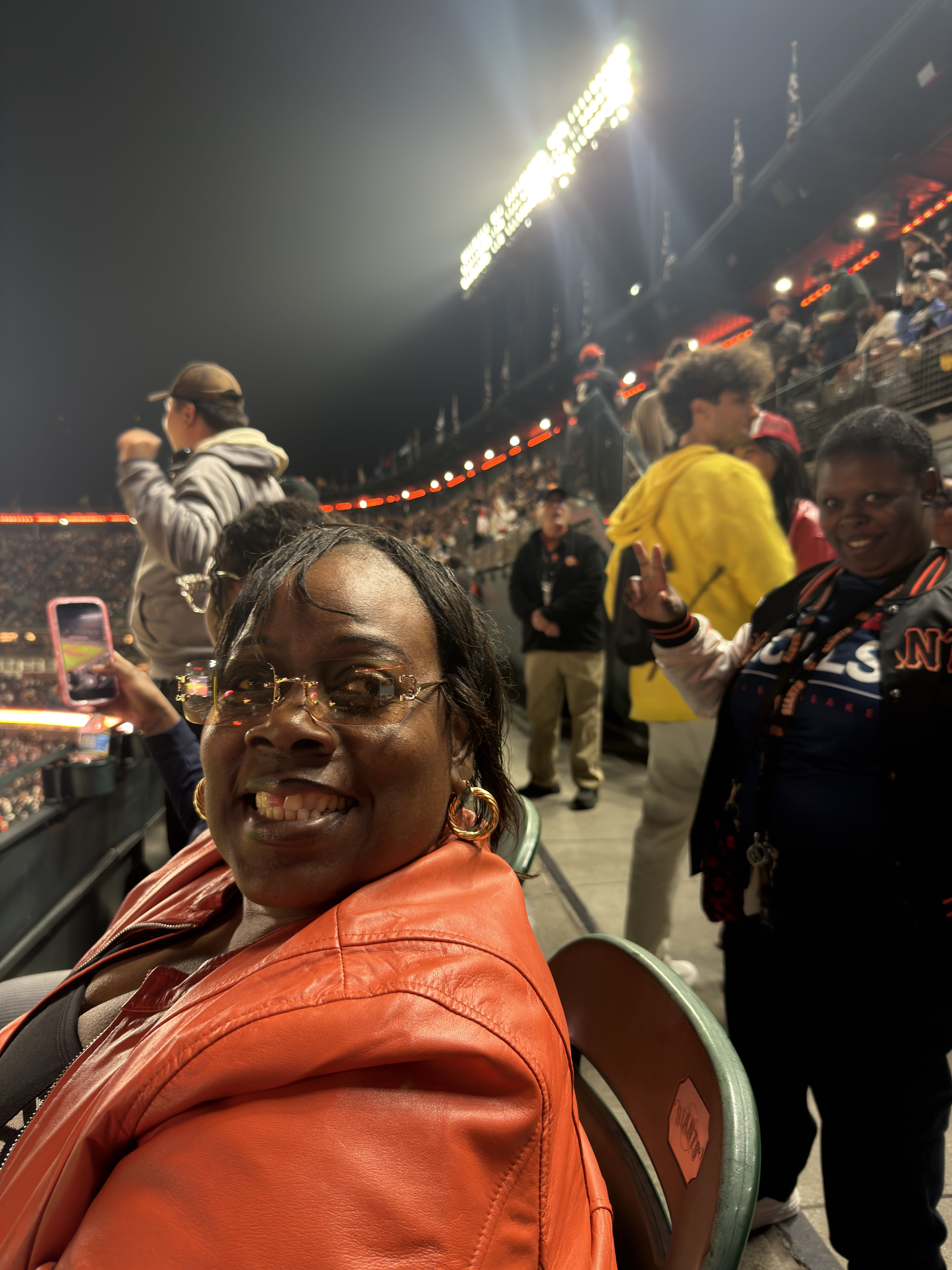 A close-up photo of a resident smiling and sitting in their seat at Oracle Park during a San Francisco Giants baseball game. They are wearing a bright orange leather jacket, gold hoop earrings, and glasses. Behind them, another resident wearing a black-and-orange Giants varsity jacket smiles and holds up peace signs for the photo. In the background, fans are seen cheering and taking pictures of the game.