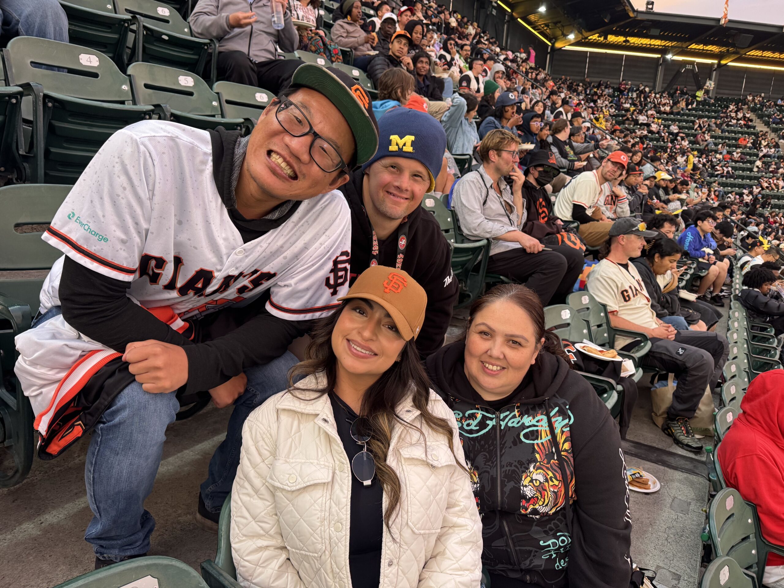 Four The Kelsey residents sitting in the stands at Oracle Park during a baseball game. Two residents are sitting in seats in the row above the other two residents. They lean into each other and smile for the picture.