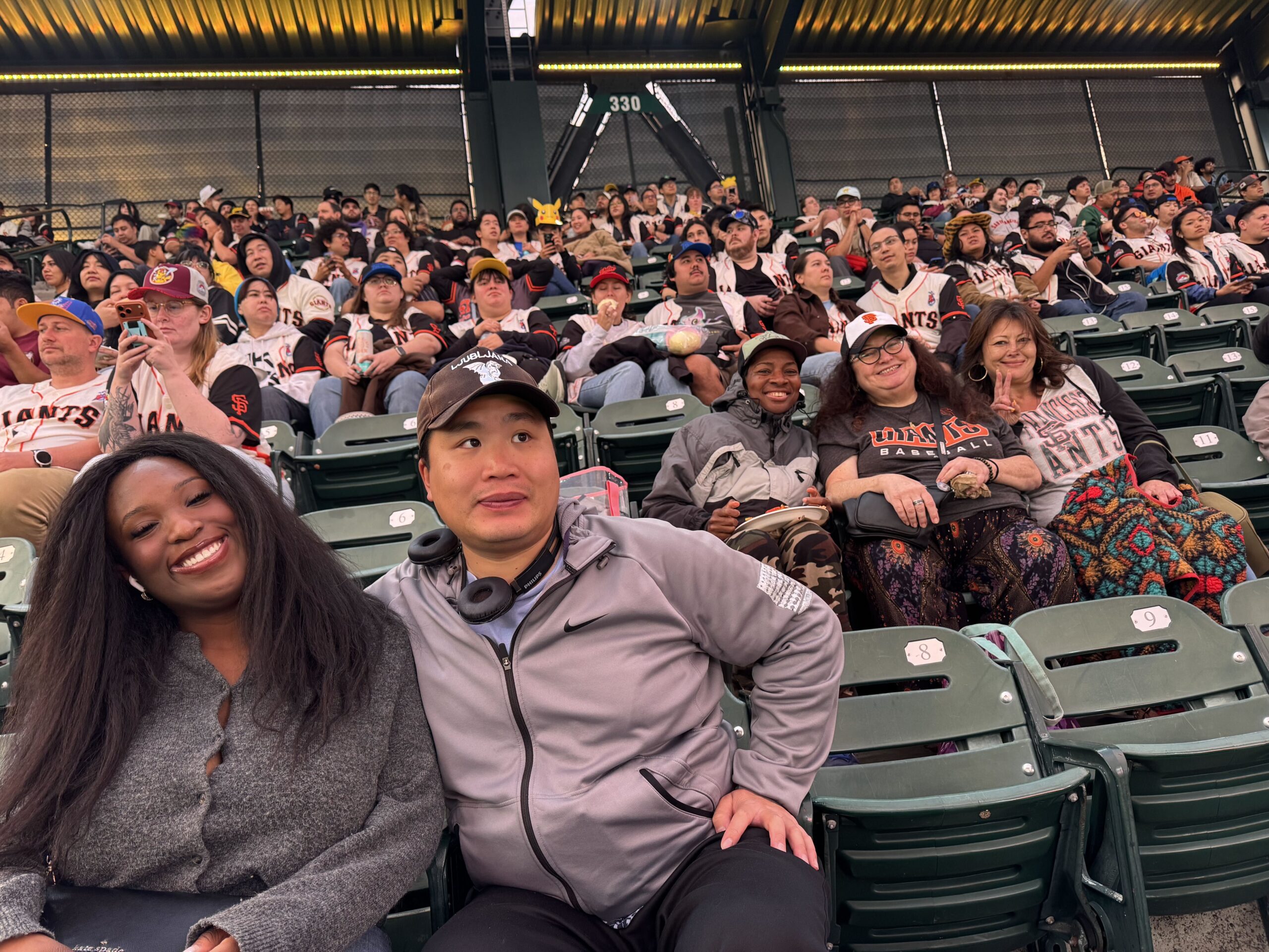 The Kelsey residents and Inclusion Concierge smile for a photo as they sit in their seats at Oracle Park for a San Francisco Giants baseball game. Two people are in the foreground of the picture, and one has their arm around the other. Sitting one row behind them are three residents leaning into each other and smiling for the photo.